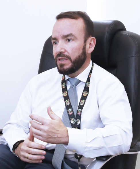 A imagem mostra um homem sentado em uma cadeira de escritório, usando camisa branca, gravata cinza e um crachá no pescoço. Ele parece estar em meio a uma conversa, gesticulando com a mão.