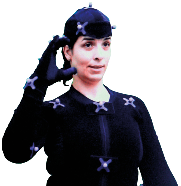 Uma mulher, com cabelo escuro preso, usa uma roupa preta com vários marcadores/sensores brancos presos para captura de movimentos. Ela levanta a mão direita, que está com uma luva preta, perto do rosto.