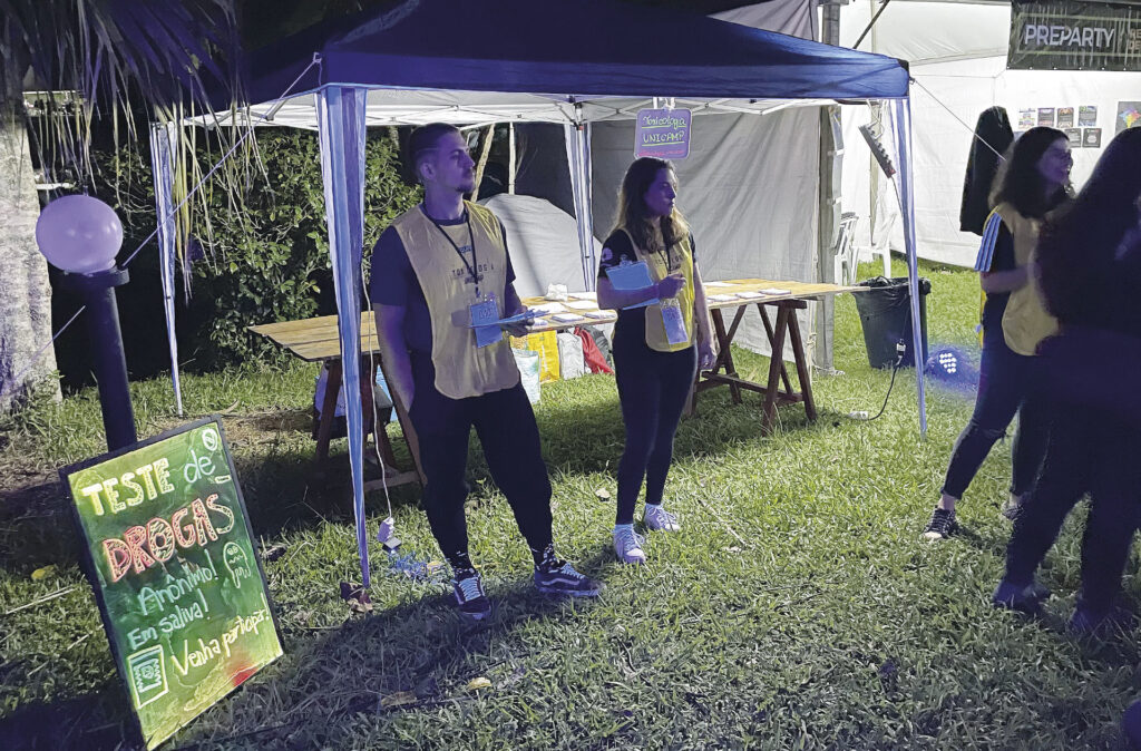 A foto retrata uma cena noturna e ao ar livre, provavelmente em um festival ou evento universitário.

Em uma área gramada, há uma tenda iluminada onde jovens, usando coletes amarelos de identificação, estão trabalhando em uma bancada. Em primeiro plano, uma pequena lousa escrita à mão anuncia um "TESTE DE DROGAS", informando que o serviço é anônimo, realizado com saliva, e convida as pessoas a participar. A atmosfera é de um evento de conscientização ou redução de danos.