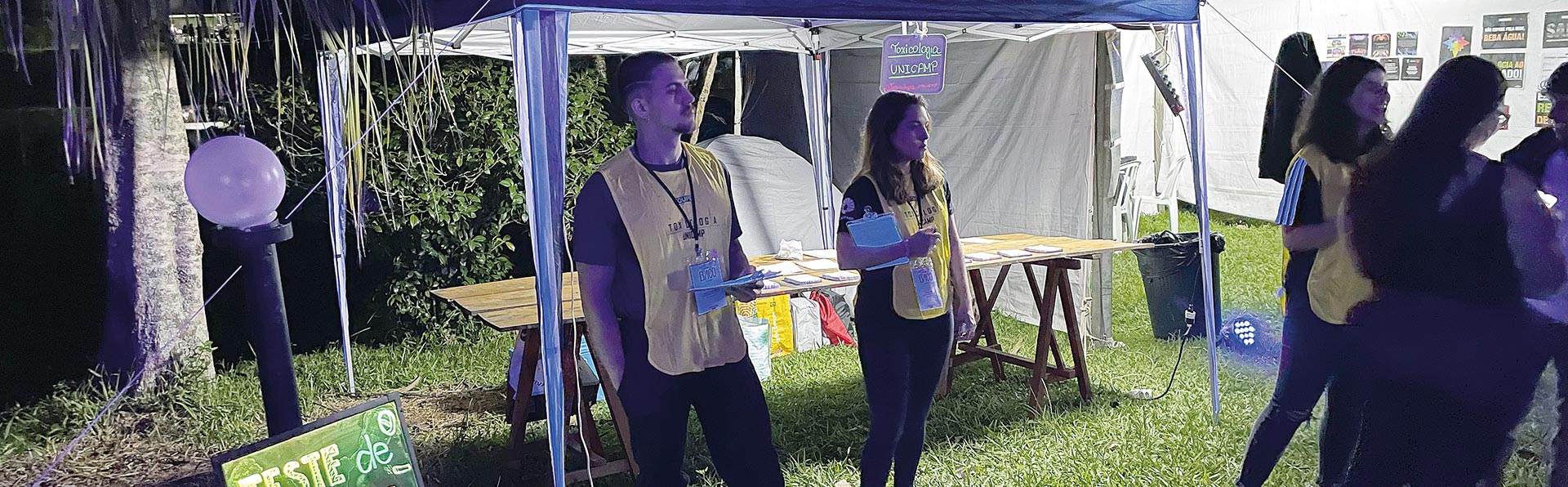 A foto retrata uma cena noturna e ao ar livre, provavelmente em um festival ou evento universitário. Em uma área gramada, há uma tenda iluminada onde jovens, usando coletes amarelos de identificação, estão trabalhando em uma bancada. Em primeiro plano, uma pequena lousa escrita à mão anuncia um "TESTE DE DROGAS", informando que o serviço é anônimo, realizado com saliva, e convida as pessoas a participar. A atmosfera é de um evento de conscientização ou redução de danos.