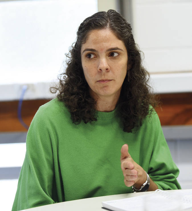 A imagem mostra uma mulher de cabelos cacheados escuros, usando uma blusa verde. Ela está sentada à mesa, com um caderno ou documento à sua frente, e parece estar falando ou explicando algo, gesticulando com a mão.