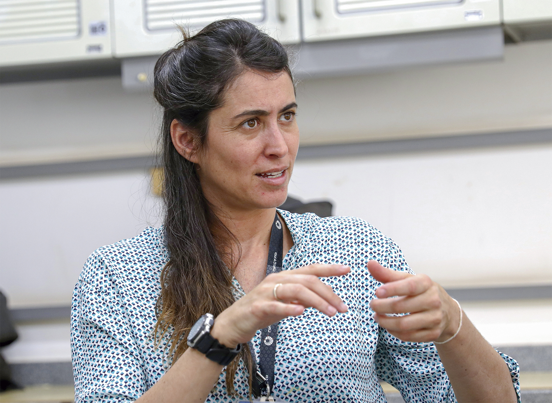 Uma mulher de pele clara e cabelos castanhos longos e ondulados, parcialmente presos para trás, é fotografada em um plano médio. Ela parece estar no meio de uma conversa, com uma expressão concentrada e a boca ligeiramente aberta, como se estivesse falando.

Ela veste uma camisa de manga curta com uma estampa pequena e densa em tons de azul ou preto sobre um fundo branco. Em seu pulso esquerdo, ela usa um relógio preto grande e um anel, e no pulso direito, uma pulseira fina e clara. Suas mãos estão à sua frente, gesticulando para enfatizar suas palavras.

O fundo está desfocado, mas parece ser um ambiente interno, como um escritório ou laboratório, com armários de cor clara visíveis atrás dela.