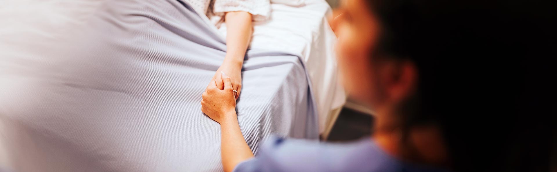 Na imagem, uma pessoa está deitada em uma cama de hospital e outra segura sua mão, transmitindo apoio e carinho.