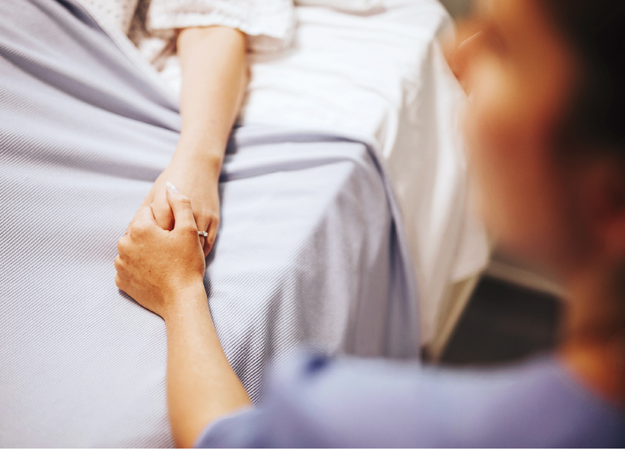 Na imagem, uma pessoa está deitada em uma cama de hospital e outra segura sua mão, transmitindo apoio e carinho.