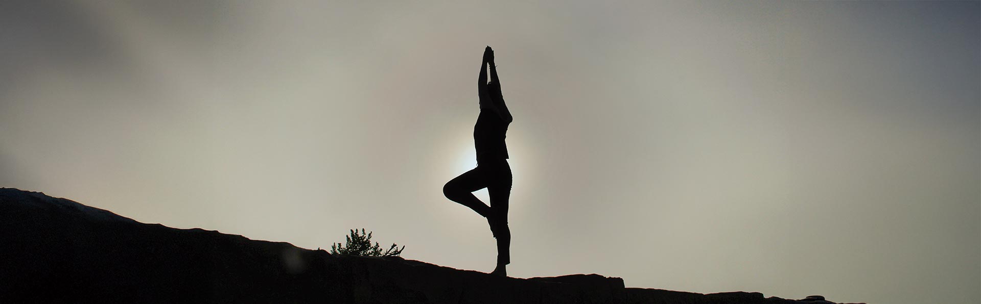 A imagem mostra a silhueta de uma pessoa em uma pose de ioga, equilibrando-se em uma perna só com os braços erguidos acima da cabeça. A figura escura se destaca contra um céu muito claro, com o sol brilhando intensamente por trás dela, criando um contorno de luz ao redor do corpo. A pessoa está em cima de uma superfície rochosa ou no topo de um morro. A cena transmite uma sensação de paz e equilíbrio.
