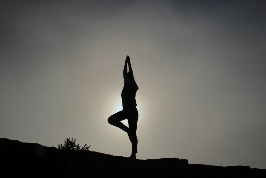 A imagem mostra a silhueta de uma pessoa em uma pose de ioga, equilibrando-se em uma perna só com os braços erguidos acima da cabeça.

A figura escura se destaca contra um céu muito claro, com o sol brilhando intensamente por trás dela, criando um contorno de luz ao redor do corpo. A pessoa está em cima de uma superfície rochosa ou no topo de um morro. A cena transmite uma sensação de paz e equilíbrio.