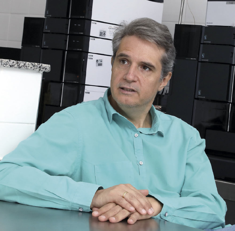 A foto retrata um homem de meia-idade com cabelos castanhos grisalhos, vestindo uma camisa social de cor verde-clara.

Ele está sentado a uma mesa com as mãos cruzadas à sua frente. Seu rosto está virado para o lado e ele parece estar no meio de uma conversa. Ao fundo, há uma grande pilha de equipamentos eletrônicos pretos, sugerindo que ele está em um ambiente de trabalho, como um laboratório ou escritório.