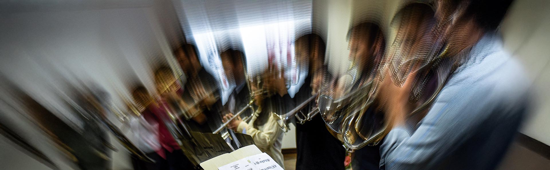 A foto captura uma apresentação musical de forma dinâmica. Em primeiro plano e em foco, há uma estante com uma partitura musical. Ao fundo, um grupo de músicos está de pé, segurando instrumentos de sopro, como trompetes. A imagem deles está com um forte efeito de desfoque de movimento, criando rastros de luz e transmitindo uma intensa sensação de energia e som.