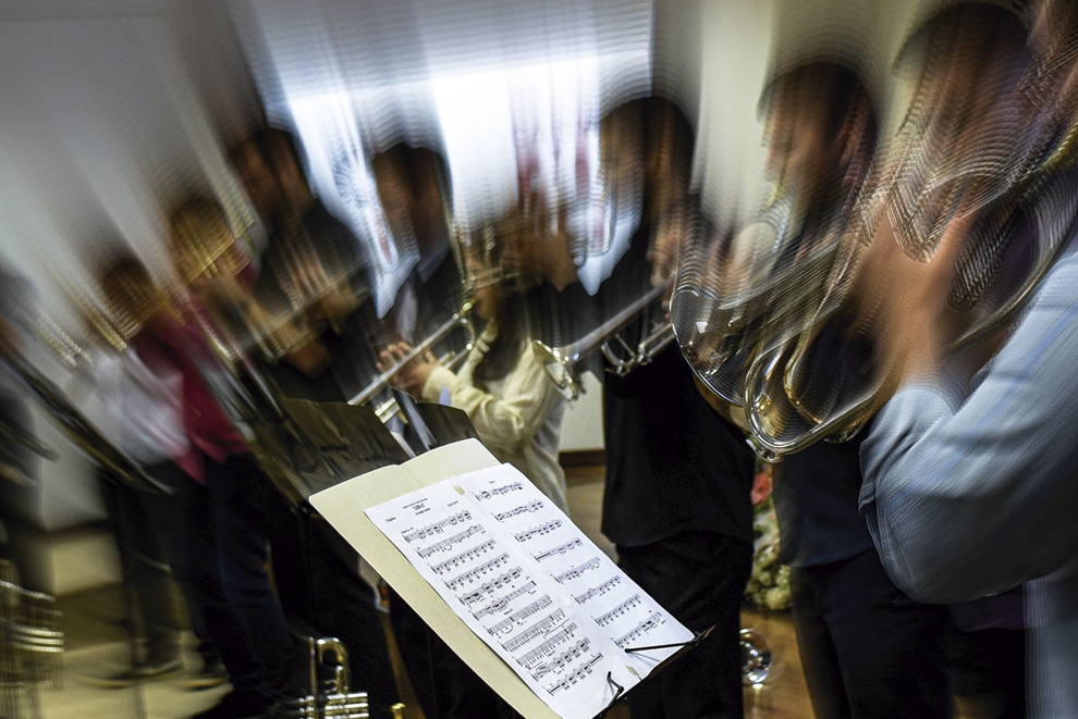A foto captura uma apresentação musical de forma dinâmica. Em primeiro plano e em foco, há uma estante com uma partitura musical.

Ao fundo, um grupo de músicos está de pé, segurando instrumentos de sopro, como trompetes. A imagem deles está com um forte efeito de desfoque de movimento, criando rastros de luz e transmitindo uma intensa sensação de energia e som.