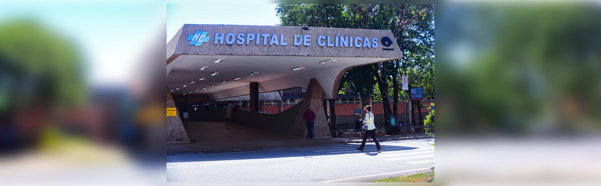 Esta é uma fotografia externa e diurna da entrada principal do Hospital de Clínicas. A imagem foi capturada em um dia ensolarado, com luz forte e sombras bem marcadas no chão. Elemento Central: O foco principal é a entrada de um grande edifício, com uma arquitetura de estilo modernista ou brutalista, caracterizada pelo uso de concreto aparente com textura granulada. Uma grande marquise se projeta sobre uma via de acesso para veículos e pedestres, criando uma área coberta. Na fachada da marquise, em letras grandes e tridimensionais de cor azul-clara, está escrito "HOSPITAL DE CLÍNICAS". À esquerda do nome, há um logotipo estilizado com as letras "HC". Pessoas na Cena: Em primeiro plano, atravessando a rua sobre uma faixa de pedestres, uma pessoa caminha da direita para a esquerda. Ela parece ser uma mulher jovem, de cabelos cacheados na altura dos ombros. Veste uma blusa de cor clara, possivelmente amarela, calças escuras e carrega uma bolsa ou mochila no ombro direito. Mais ao fundo, sob a área coberta da entrada, podem ser vistas mais duas pessoas em pé, de forma menos nítida. Uma delas usa uma camisa de cor avermelhada. Ambiente e Entorno: O hospital está localizado em uma área bastante arborizada. Ao fundo e nas laterais da entrada, há várias árvores altas com folhagem verde e densa, que recebem a luz direta do sol. A iluminação geral é clara e vibrante, típica de um dia de céu aberto. A rua em primeiro plano é de asfalto escuro.