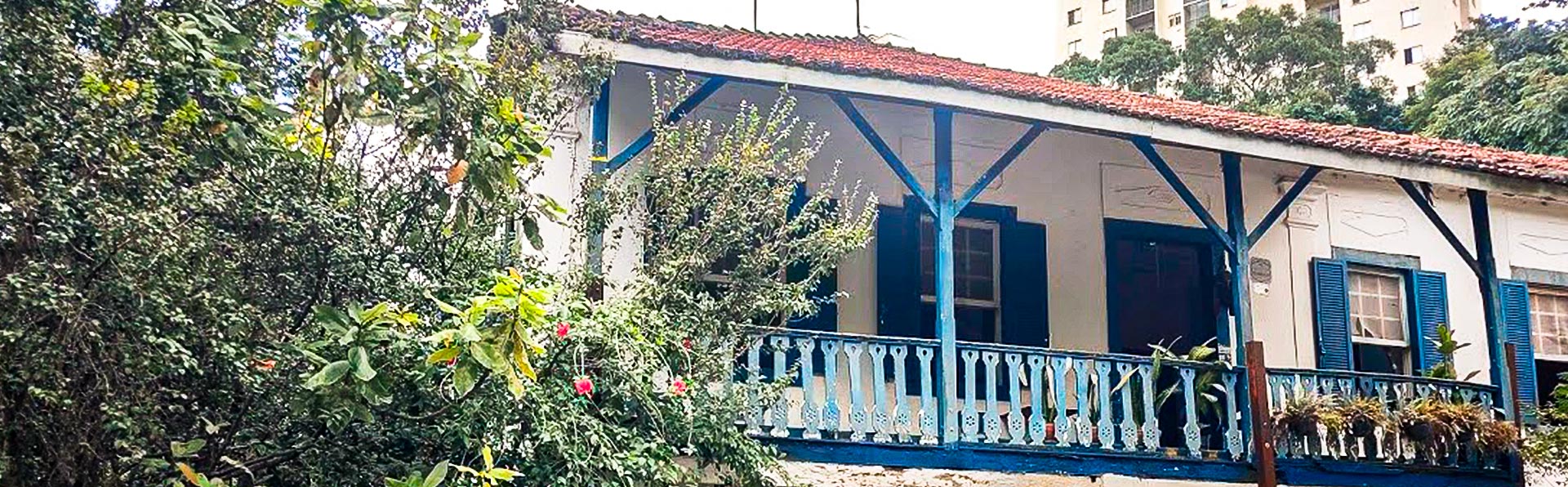 A foto mostra a fachada de uma casa antiga, estilo colonial. As paredes são brancas, com uma varanda que tem a grade e as vigas de madeira pintadas de azul. O telhado é de telhas vermelhas de barro. A casa é cercada por um jardim verde e denso e, ao fundo, é possível ver a parte de cima de prédios modernos.
