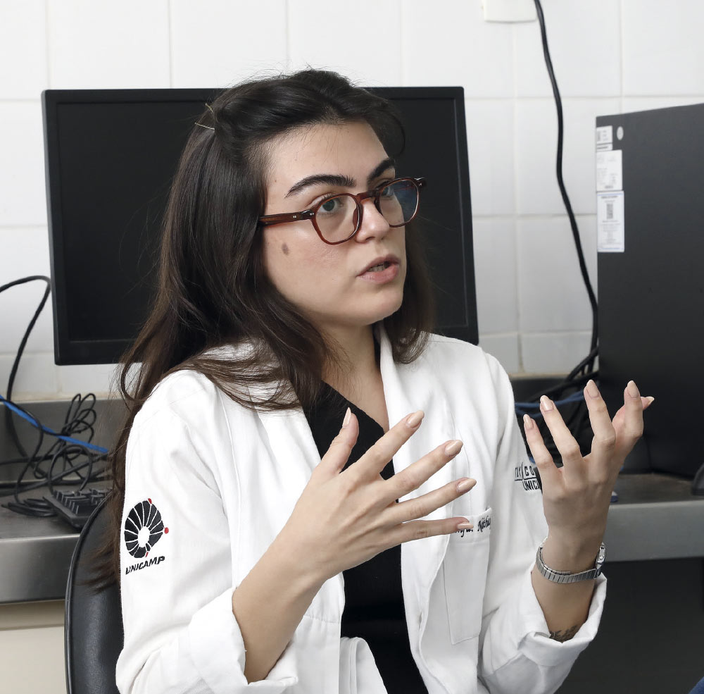 A foto retrata uma mulher jovem de cabelos longos e escuros, usando óculos de armação marrom. Ela está em um ambiente que parece ser um laboratório ou escritório, vestindo um jaleco branco sobre uma blusa preta.

Sentada, ela gesticula com as duas mãos à sua frente enquanto fala, com uma expressão concentrada, como se estivesse explicando algo importante. Ao fundo, é possível ver monitores de computador e outros equipamentos.