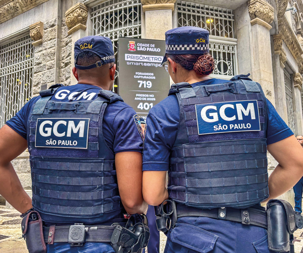 Dois guardas municipais de São Paulo, um homem e uma mulher, estão de costas, olhando para um painel digital. Ambos usam uniformes e coletes azuis com o emblema da GCM São Paulo. No painel, aparece o título "Prisômetro" e o número de prisões realizadas pela guarda.