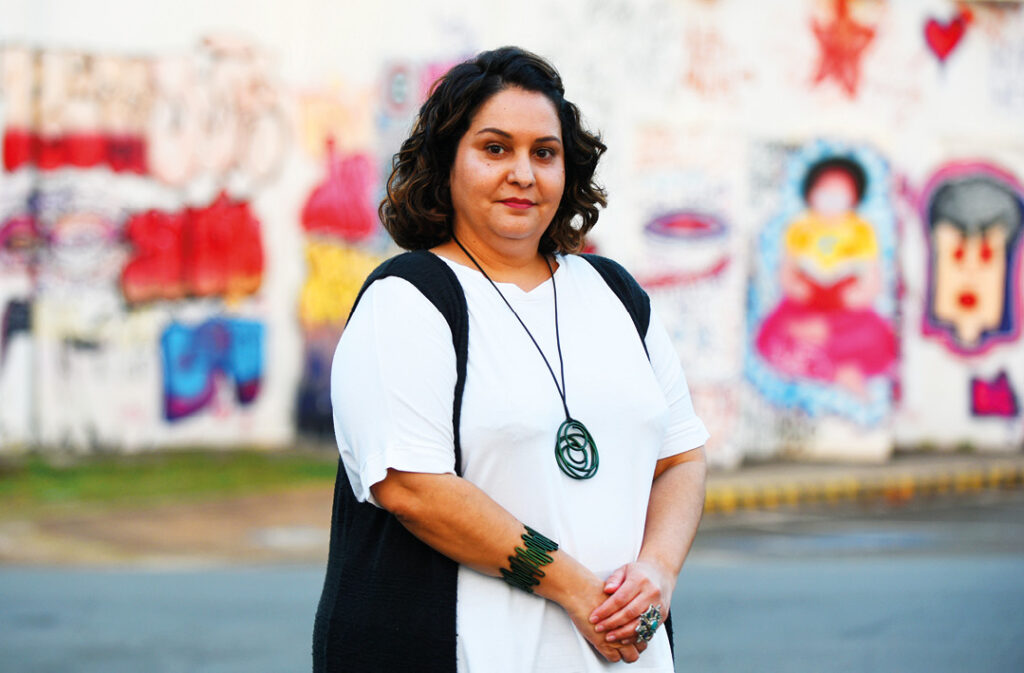 Uma mulher de cabelo escuro, com uma camiseta branca e um colete preto, está em frente a uma parede com grafite colorido.
