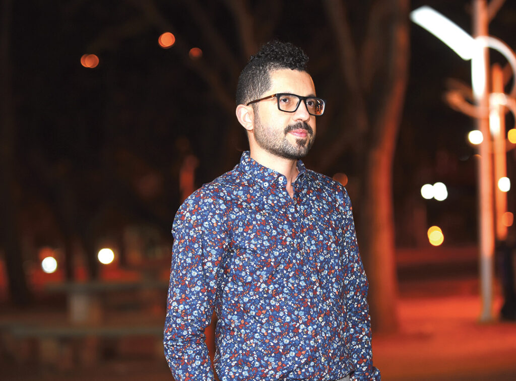 Um homem com barba e óculos usa uma camisa floral azul. Ele está em um parque à noite. O fundo está escuro e borrado, com luzes de postes.