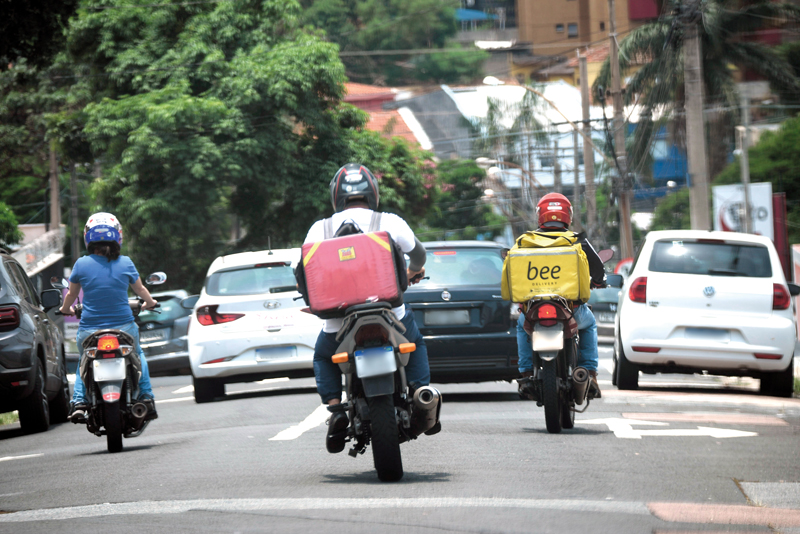 A imagem mostra uma rua movimentada com vários veículos e três motociclistas em destaque. Os motociclistas estão de capacete e vestem roupas casuais; dois deles carregam mochilas grandes de entrega nas costas — uma vermelha e outra amarela. Há carros à frente e ao lado deles, e o cenário urbano ao fundo inclui árvores, postes e prédios. A cena ocorre durante o dia, sob luz natural.