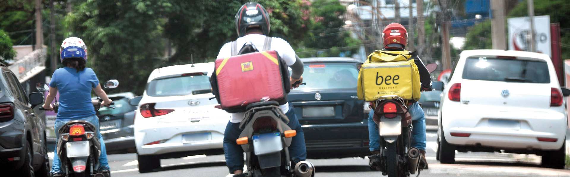 A imagem mostra uma rua movimentada com vários veículos e três motociclistas em destaque. Os motociclistas estão de capacete e vestem roupas casuais; dois deles carregam mochilas grandes de entrega nas costas — uma vermelha e outra amarela. Há carros à frente e ao lado deles, e o cenário urbano ao fundo inclui árvores, postes e prédios. A cena ocorre durante o dia, sob luz natural.