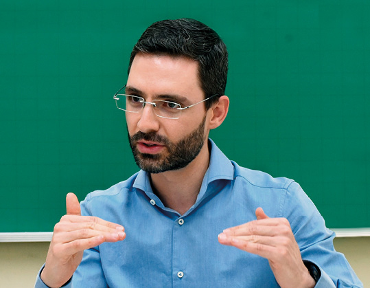 Um homem branco, de barba e óculos, com uma camisa azul-clara, aparece em um retrato de close-up. Ele gesticula com as duas mãos abertas, em frente a um fundo liso de cor verde escura, com uma expressão focada enquanto explica algo.