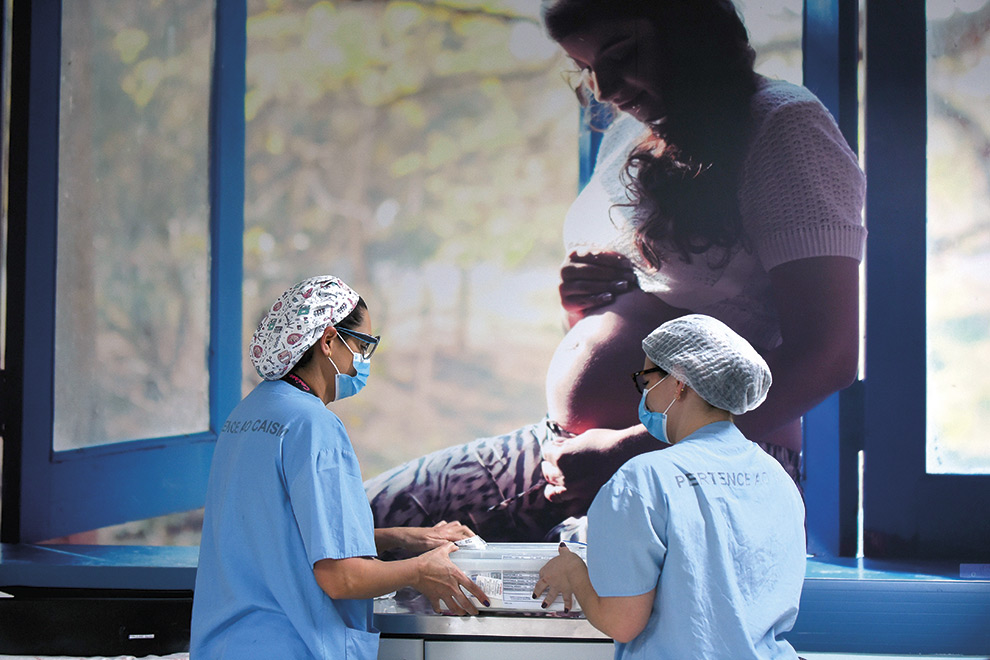 A imagem retrata duas profissionais de saúde, vistas predominantemente de costas, em um ambiente clínico ou hospitalar. Elas vestem uniformes cirúrgicos azuis, toucas, máscaras faciais e óculos de proteção. As duas estão em pé, próximas uma da outra, manuseando com cuidado um recipiente plástico transparente sobre uma superfície de aço inoxidável. Ao fundo, ocupando quase toda a parede, há um grande painel fotográfico com a imagem de uma mulher grávida, de perfil, acariciando a barriga com as mãos em um gesto de ternura. A cena contrasta o ambiente técnico e asséptico do hospital com a imagem calorosa e humana da maternidade.
