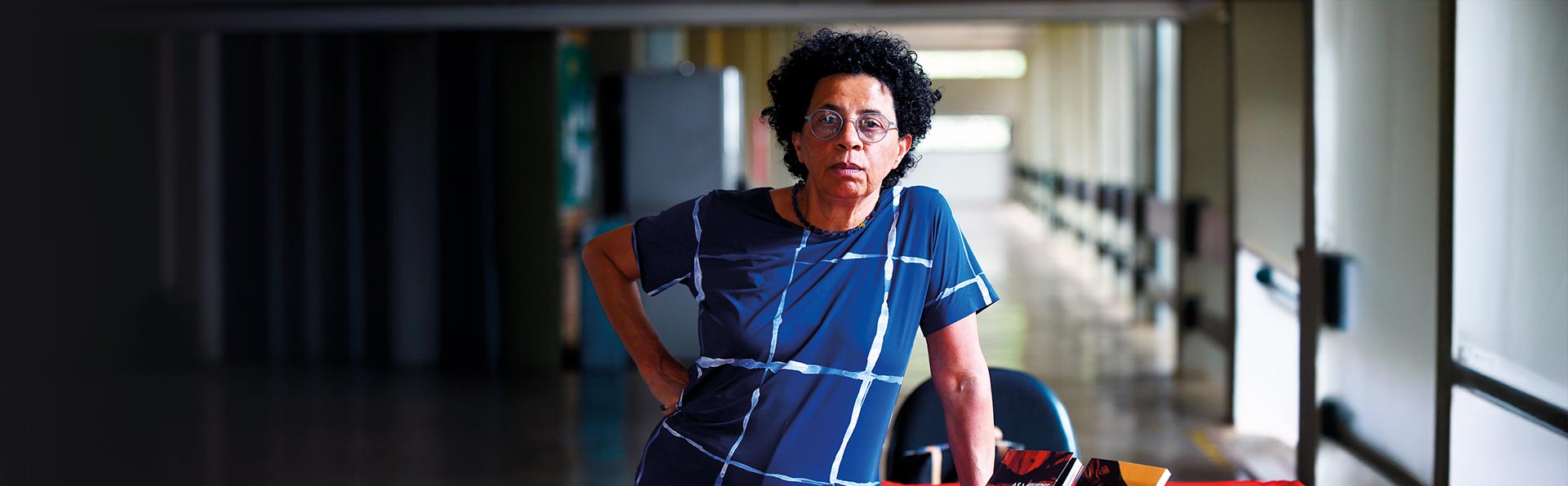 Em uma foto panorâmica horizontal, uma mulher de cabelo escuro, curto e cacheado está posicionada do lado direito da imagem. Ela usa óculos redondos e uma blusa azul-escura com um padrão de linhas brancas que se cruzam. Ela está de pé, com uma das mãos na cintura, olhando diretamente para a câmera com uma expressão séria. À sua frente, no canto inferior direito, há alguns livros sobre uma superfície. O fundo atrás dela é um corredor; o lado esquerdo da imagem é muito escuro e sombrio, enquanto o lado direito do corredor é mais claro e mostra uma série de colunas ou aberturas.