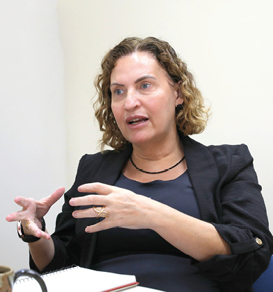 A imagem mostra uma mulher sentada à mesa, falando e gesticulando com as mãos. Ela tem cabelo castanho claro, cacheado e na altura dos ombros. Usa um blazer preto sobre uma blusa escura e um colar simples. À sua frente há um caderno de anotações. O ambiente é claro e parece ser um escritório ou sala de reunião.