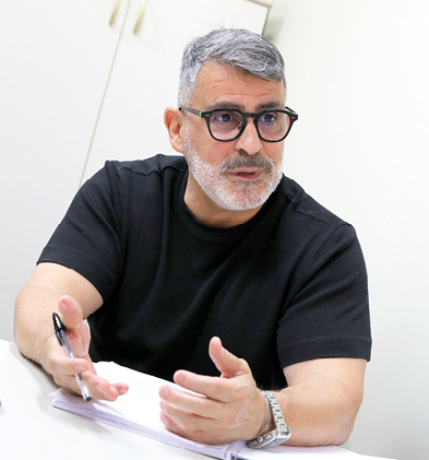 A imagem mostra um homem de meia-idade sentado à mesa, falando e gesticulando com as mãos. Ele tem barba grisalha, cabelo curto também grisalho e usa óculos de armação escura. Está vestido com uma camiseta preta e um relógio prateado. Diante dele há um caderno ou bloco de anotações e uma caneta. O ambiente parece ser um escritório, com paredes claras e armários ao fundo.