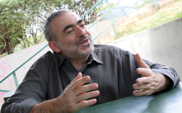 A imagem mostra um homem sentado ao ar livre, falando e gesticulando com as mãos. Ele tem barba grisalha curta e cabelo também grisalho, veste uma camisa escura de mangas compridas. Está encostado em uma mesa verde, e ao fundo há uma mureta, escadas e vegetação. A expressão do homem é simpática, como se estivesse em uma conversa animada.