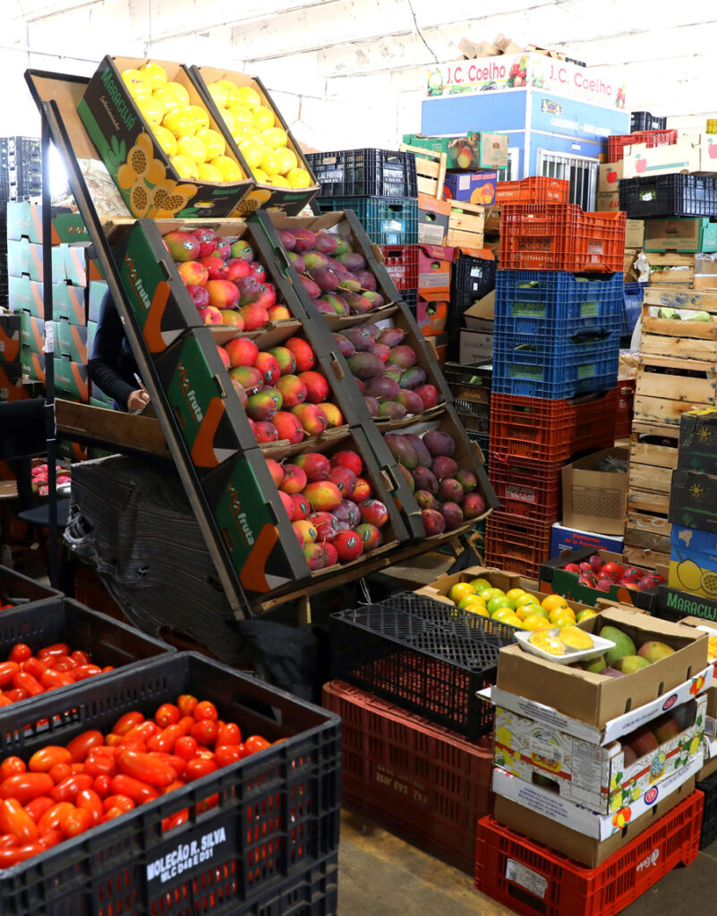 A imagem mostra o interior de um mercado ou armazém de frutas e verduras. Há várias caixas plásticas empilhadas, cheias de produtos coloridos. À esquerda, há muitas caixas pretas com tomates vermelhos. No centro, há uma estrutura inclinada com caixas de mangas e limões. À direita, há mais caixas com frutas variadas, como mangas e limões, e outras empilhadas ao fundo. O ambiente parece movimentado, cheio de mercadorias e cores vivas.