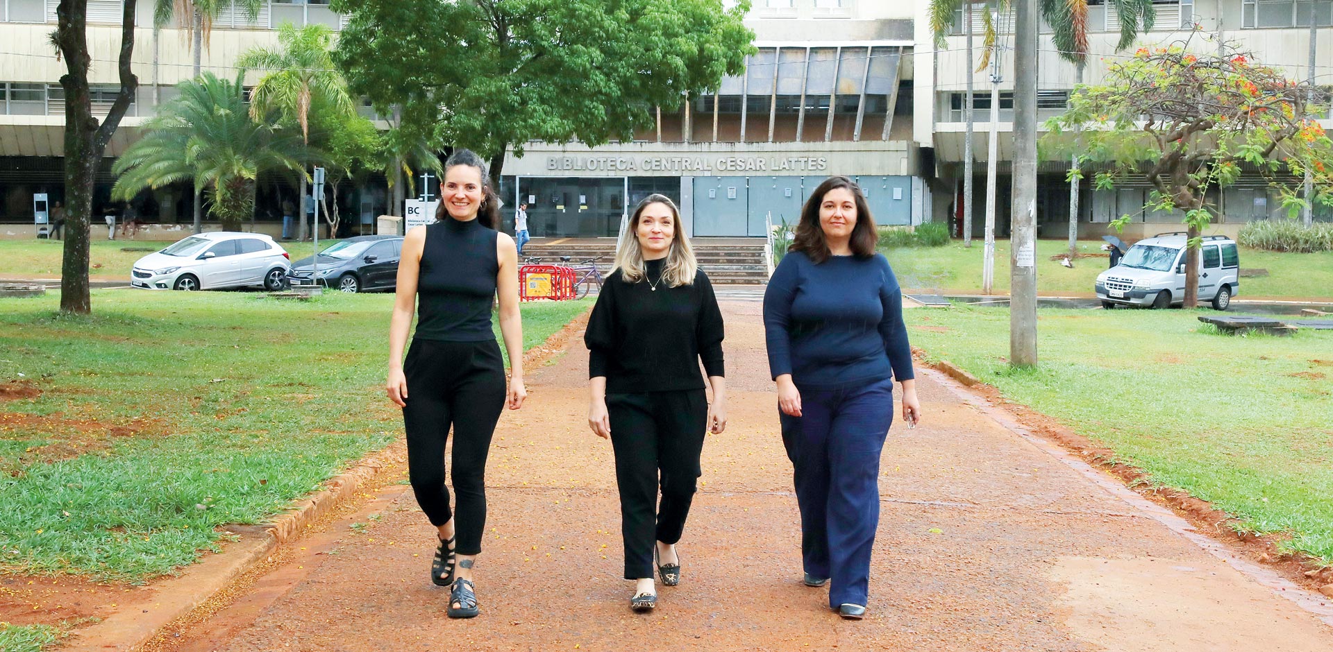 Três mulheres adultas, vestindo roupas escuras, caminham juntas em um caminho de campus de universidade. Elas avançam em direção a quem observa a foto, tendo como fundo o prédio principal da Biblioteca Central Cesar Lattes, cercado por gramado e árvores.