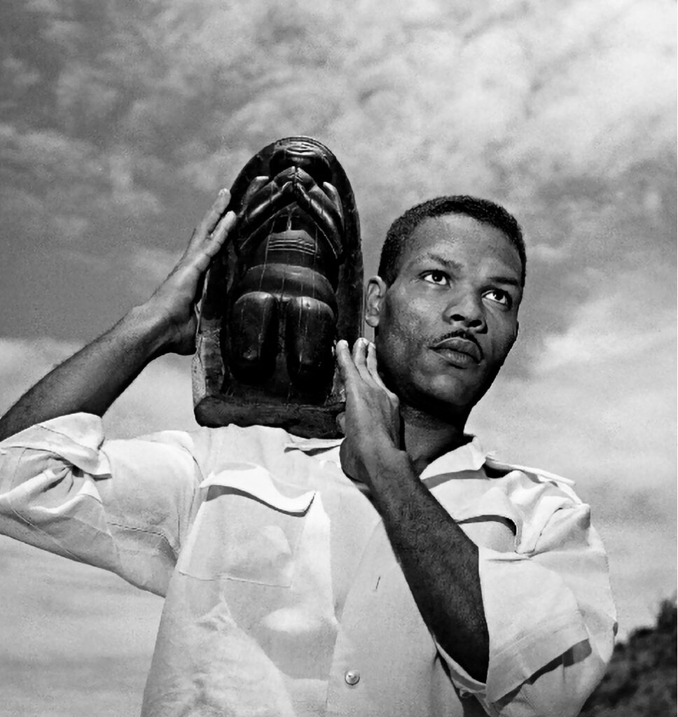 Esta é uma fotografia impactante em preto e branco, tirada de um ângulo baixo. A imagem mostra um homem negro, com um bigode fino, vestindo uma camisa clara de mangas dobradas. Ele carrega uma grande e pesada escultura de madeira em seu ombro, segurando-a com as duas mãos. A escultura representa uma figura ou rosto estilizado com as mãos sobre a boca. O homem olha para cima e para o lado, com uma expressão séria e determinada. O fundo é um céu com nuvens, o que confere à cena uma sensação de força e monumentalidade.