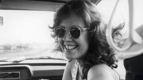 Esta é uma fotografia em preto e branco do rosto de uma mulher sorrindo, tirada de dentro de um carro. Ela tem cabelo na altura dos ombros, com ondas, e usa óculos de sol redondos. Ela está olhando para a câmera com um sorriso largo e alegre. O interior do carro, incluindo parte do painel e o espelho retrovisor, é visível ao fundo. A imagem tem uma aparência clássica e espontânea.