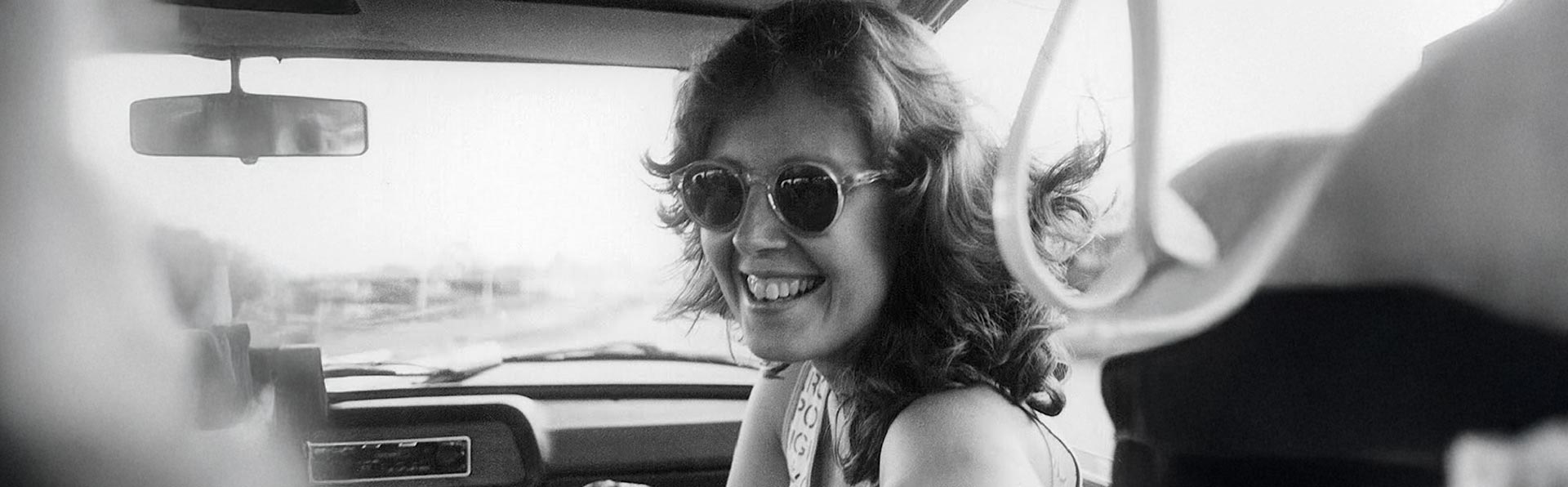 Esta é uma fotografia em preto e branco do rosto de uma mulher sorrindo, tirada de dentro de um carro. Ela tem cabelo na altura dos ombros, com ondas, e usa óculos de sol redondos. Ela está olhando para a câmera com um sorriso largo e alegre. O interior do carro, incluindo parte do painel e o espelho retrovisor, é visível ao fundo. A imagem tem uma aparência clássica e espontânea.