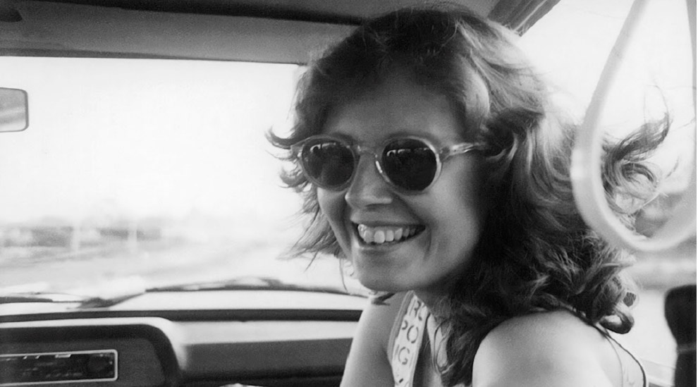 Esta é uma fotografia em preto e branco do rosto de uma mulher sorrindo, tirada de dentro de um carro. Ela tem cabelo na altura dos ombros, com ondas, e usa óculos de sol redondos. Ela está olhando para a câmera com um sorriso largo e alegre. O interior do carro, incluindo parte do painel e o espelho retrovisor, é visível ao fundo. A imagem tem uma aparência clássica e espontânea.