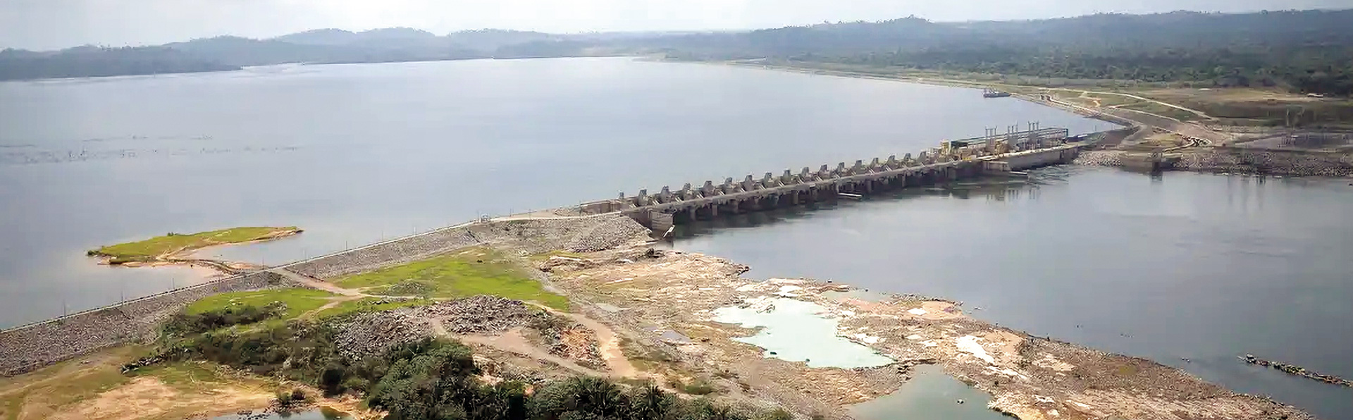 Uma foto aérea mostra uma barragem construída sobre um grande rio ou lago. No centro, a estrutura longa de concreto e terra divide o vasto reservatório azul-acinzentado no fundo. Em primeiro plano, há terra com vegetação e solo seco, com floresta verde ao redor, sob um céu claro.