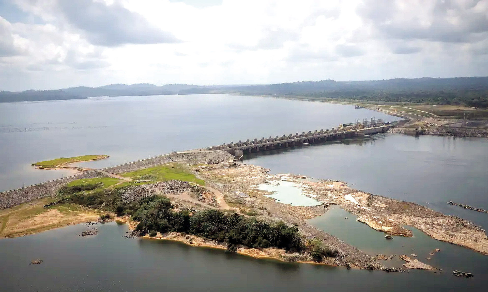 Uma foto aérea mostra uma barragem construída sobre um grande rio ou lago. No centro, a estrutura longa de concreto e terra divide o vasto reservatório azul-acinzentado no fundo. Em primeiro plano, há terra com vegetação e solo seco, com floresta verde ao redor, sob um céu claro.