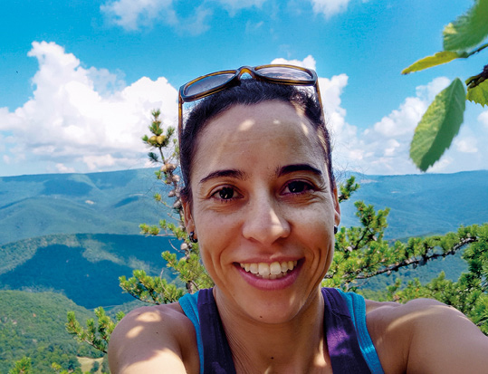 Uma mulher sorridente faz uma selfie ao ar livre. Ela está em primeiro plano, contra um vasto cenário de montanhas cobertas de mata verde e um céu azul com nuvens brancas.