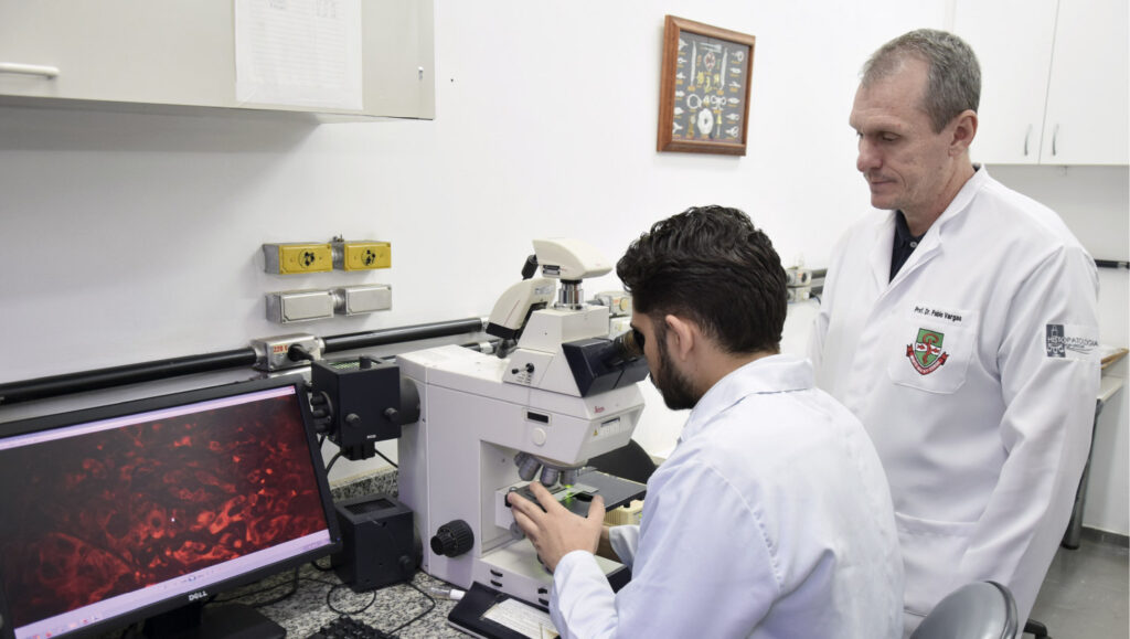Esta imagem retrata uma cena dentro de um laboratório de pesquisa ou análise. Em primeiro plano, um homem jovem com cabelo escuro e barba está sentado, vestindo um jaleco branco e olhando atentamente através das lentes de um grande e moderno microscópio. Ao seu lado, em pé, um homem mais velho, também de jaleco, o observa, parecendo supervisionar ou orientar o trabalho. À esquerda, um monitor de computador exibe uma imagem de microscopia com células ou tecidos em tons de vermelho brilhante. O ambiente é bem iluminado e funcional, típico de um espaço científico.