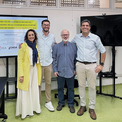 Esta é uma fotografia de um grupo de quatro pessoas — uma mulher e três homens — posando sorridentes em um ambiente que parece ser uma sala de universidade ou um auditório.
Da esquerda para a direita, vemos uma mulher com cabelos cacheados escuros, vestindo um blazer amarelo sobre um vestido claro. Ao seu lado, está um homem alto de óculos e camisa social. No centro, um homem mais velho, calvo, com barba e óculos. Na extrema direita, outro homem alto sorri, vestindo uma camisa de botões clara.
Eles estão em pé em frente a uma tela de projeção que exibe um slide de apresentação com o título de uma "Defesa de Dissertação". A atmosfera é positiva e de celebração, provavelmente registrando o final de uma banca acadêmica.