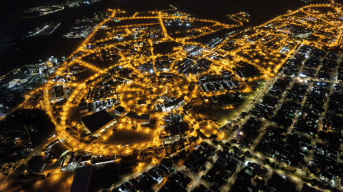Foto aérea noturna mostrando campus da Unicamp vista de cima. A imagem é dominada pelas luzes amarelas brilhantes das ruas e edifícios, que traçam o formato da área contra a escuridão da noite.