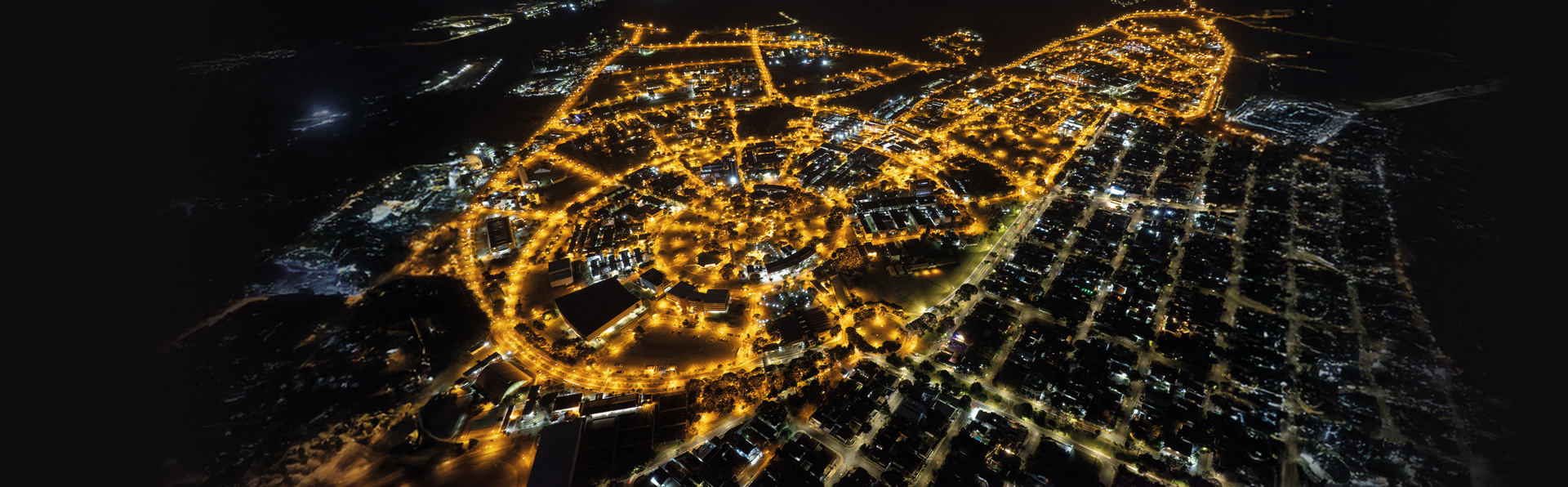 Foto aérea noturna mostrando campus da Unicamp vista de cima. A imagem é dominada pelas luzes amarelas brilhantes das ruas e edifícios, que traçam o formato da área contra a escuridão da noite.