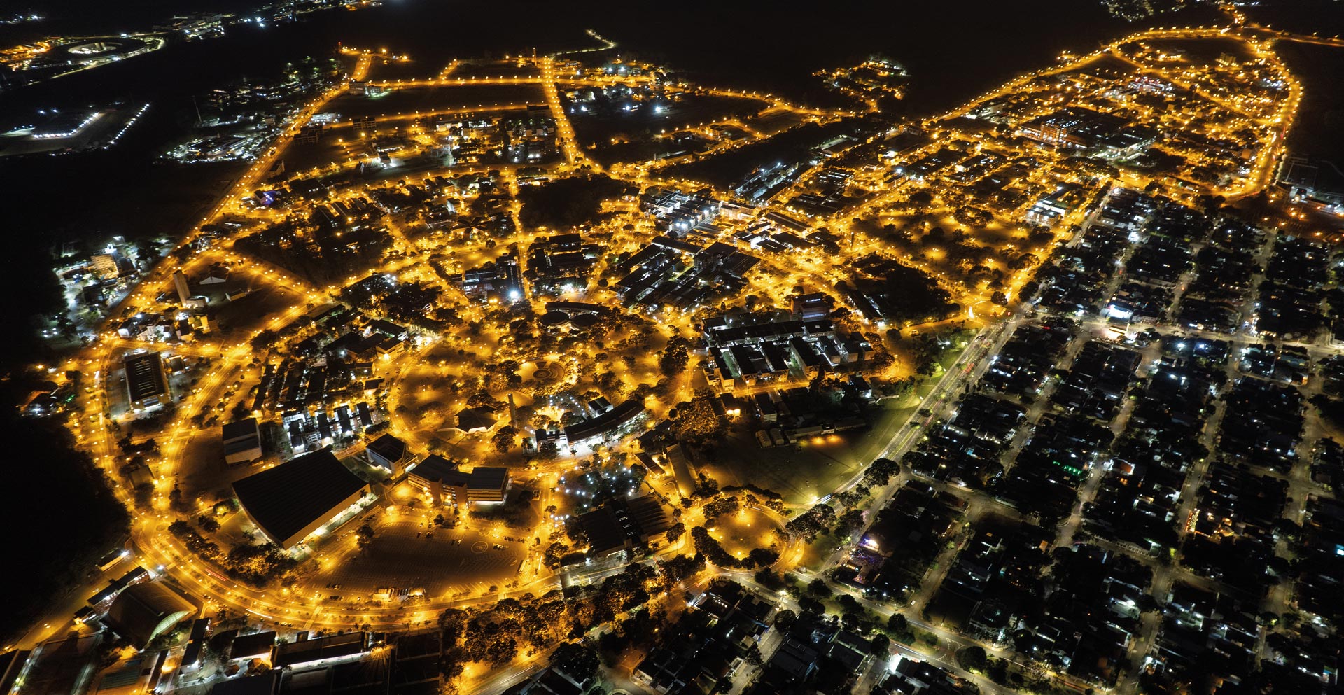 Foto aérea noturna mostrando campus da Unicamp vista de cima. A imagem é dominada pelas luzes amarelas brilhantes das ruas e edifícios, que traçam o formato da área contra a escuridão da noite.
