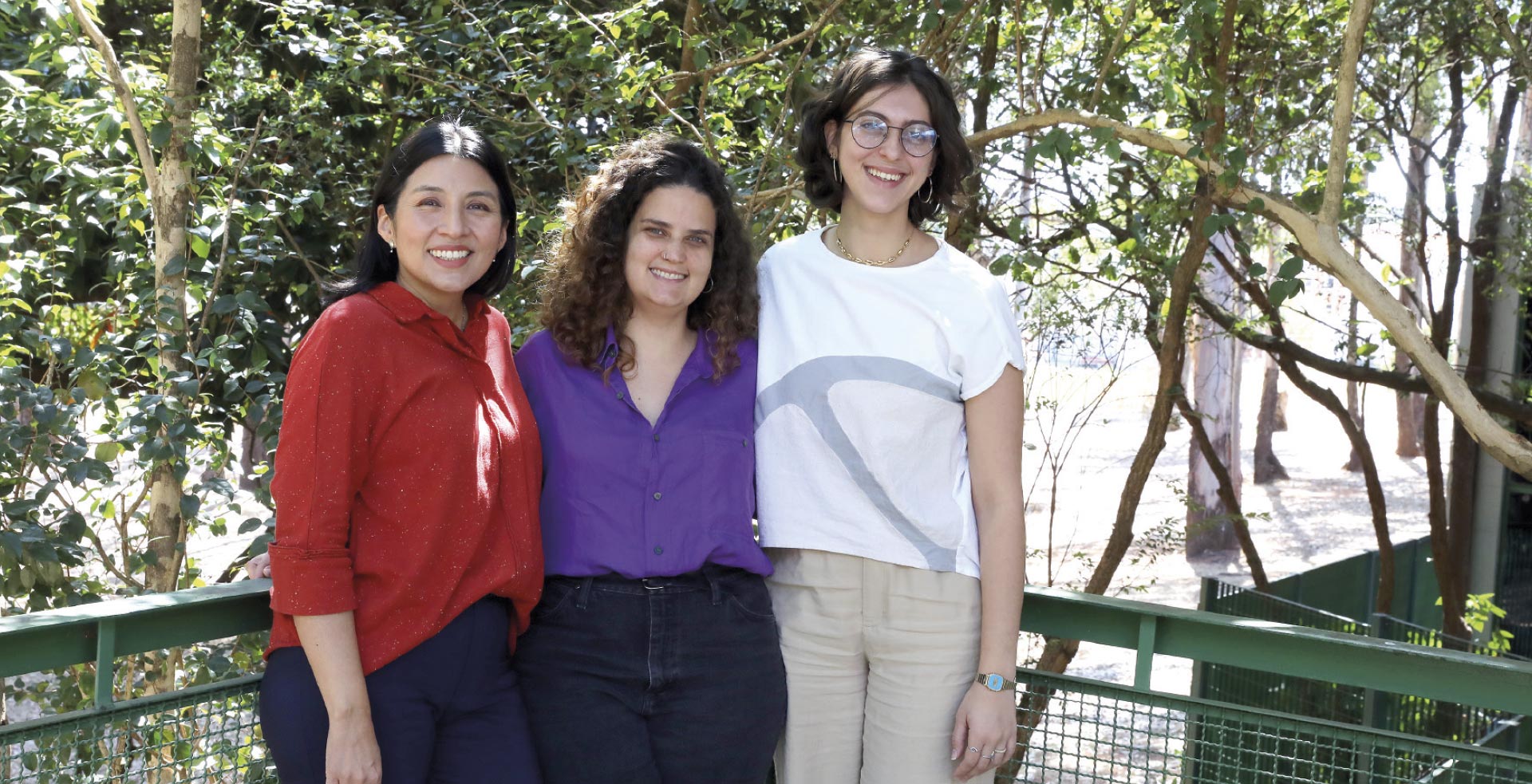 Esta é uma fotografia de três mulheres jovens, posando juntas, lado a lado, em um ambiente externo e ensolarado, com muitas árvores e folhagem verde ao fundo.
A mulher à esquerda tem cabelos longos, escuros e lisos, veste uma blusa vermelha e sorri calorosamente para a câmera. A mulher no centro, entre as outras duas, tem cabelos cacheados e volumosos, e veste uma camisa de botões de cor roxa. À direita está a mulher mais alta do grupo; ela tem cabelo curto e ondulado, usa óculos e sorri abertamente, vestindo uma camiseta branca com um detalhe em cinza e calças de cor clara.
Elas estão em pé em uma passarela ou varanda com um guarda-corpo verde, e a atmosfera geral da imagem é de amizade, descontração e alegria.