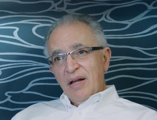 A imagem mostra um homem de meia-idade com cabelos grisalhos e óculos de armação escura. Ele veste uma camisa social branca e parece estar falando ou sendo entrevistado. Ao fundo, há uma parede azul com um desenho em linhas brancas que lembra formas orgânicas ou ondas.