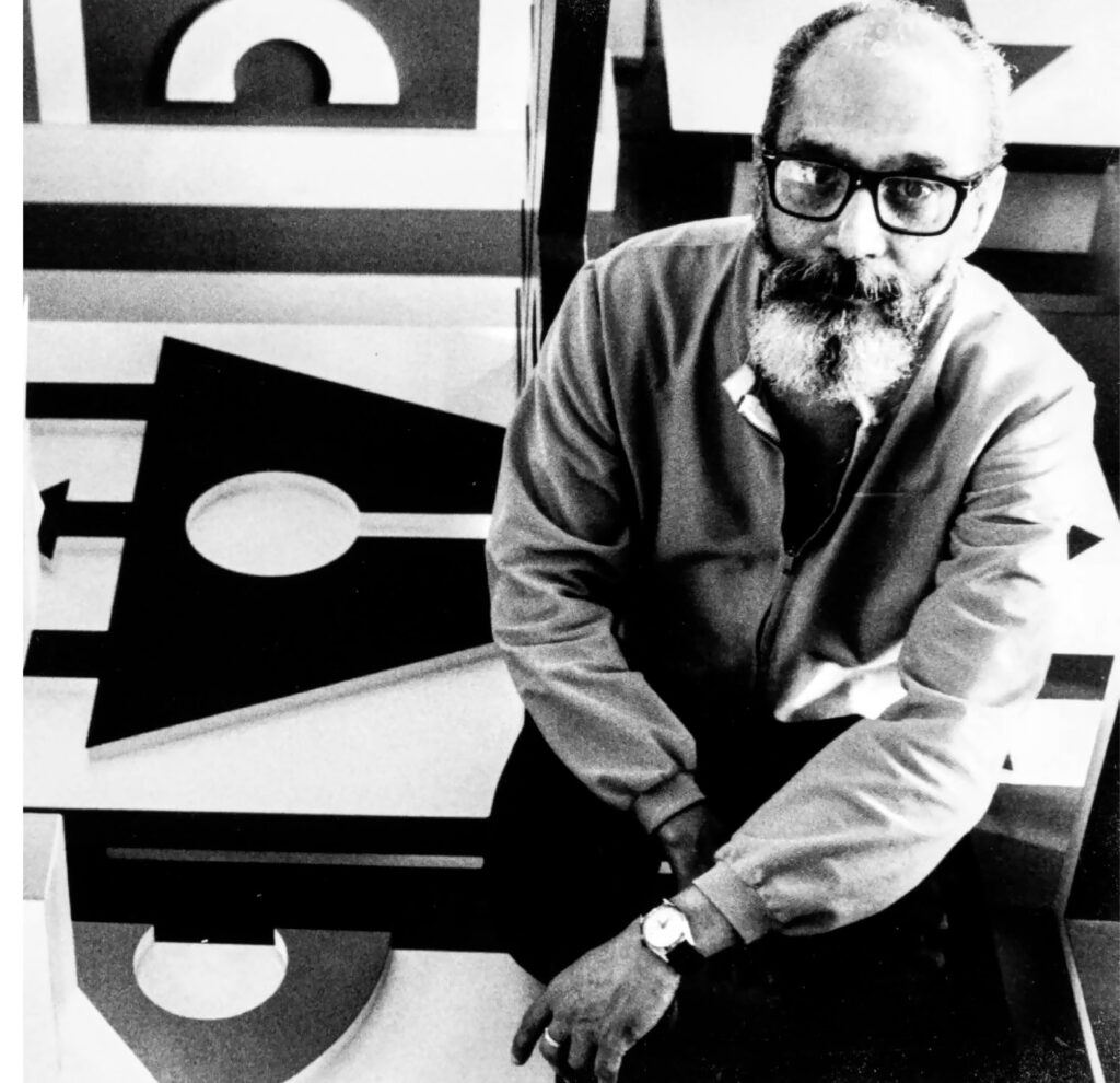 Esta é uma fotografia em preto e branco que retrata um homem de meia-idade a idoso, com uma barba cheia, bigode e óculos de armação escura. Ele está calvo no topo da cabeça, com cabelo dos lados. O homem está agachado ou sentado, olhando diretamente para a câmera com uma expressão séria e contemplativa. Ele veste uma jaqueta com zíper e, ao seu redor, há uma obra de arte abstrata com formas geométricas ousadas, como listras grossas, círculos vazados e outras figuras em preto, branco e tons de cinza.