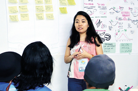 Uma mulher, com cabelo escuro e comprido, está em pé em um ambiente interno, olhando para a esquerda enquanto se dirige a um grupo de pessoas, das quais apenas as nucas de duas delas são visíveis em primeiro plano. Ela veste uma blusa clara estampada. Atrás dela, uma parede branca está coberta por post-its amarelos e um cartaz grande com desenhos e textos feitos à mão sobre temas ambientais, indicando um momento de apresentação ou debate.
