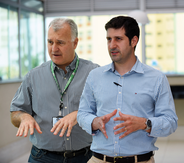 A imagem mostra dois homens em pé, conversando em um ambiente interno bem iluminado. Ambos usam camisas sociais listradas e estão gesticulando com as mãos enquanto falam. O homem à esquerda tem cabelos grisalhos e usa um crachá com cordão verde no pescoço. O homem à direita tem cabelos escuros curtos e usa um relógio no pulso. Ao fundo, há janelas amplas e luz natural entrando no local.