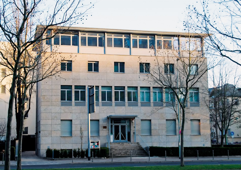 A imagem mostra a fachada de um prédio de três andares, com revestimento em pedra clara e janelas retangulares alinhadas. Há uma entrada central com escadas que levam até uma porta de vidro com cobertura metálica. À frente do edifício há uma calçada, algumas árvores sem folhas e um gramado estreito. O céu está claro, indicando um dia de sol ou fim de tarde.