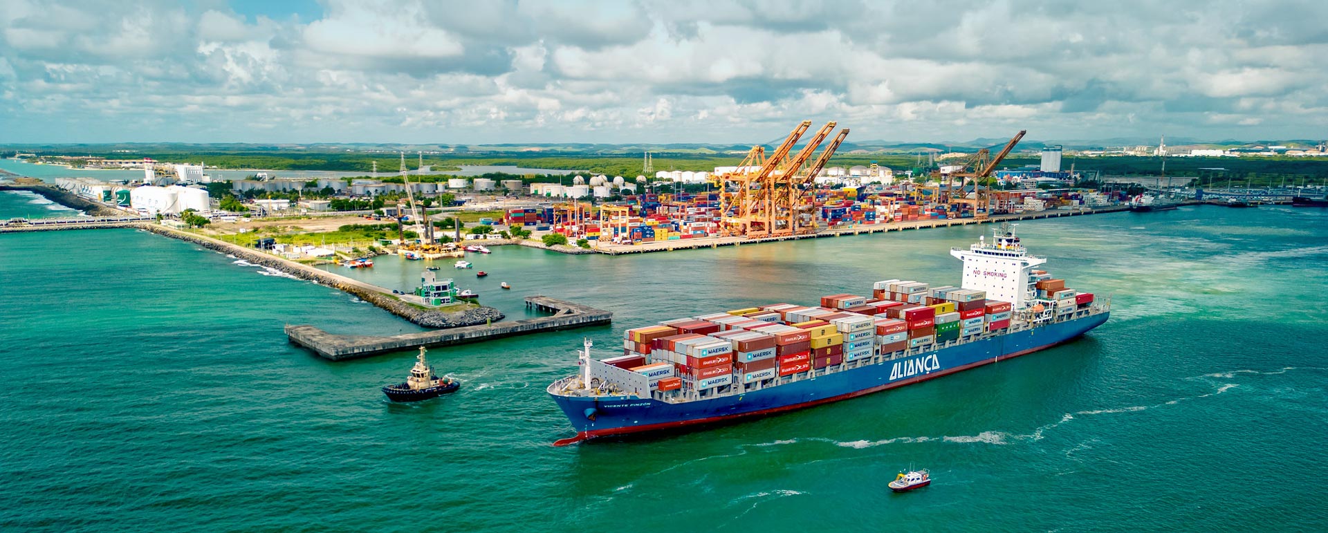 Uma foto aérea mostra um grande navio porta-contêineres azul e vermelho, carregado com contêineres coloridos, navegando em águas verde-esmeralda em primeiro plano. Ao fundo, um porto industrial está repleto de contêineres empilhados e várias grandes gruas amarelas sob um céu parcialmente nublado.