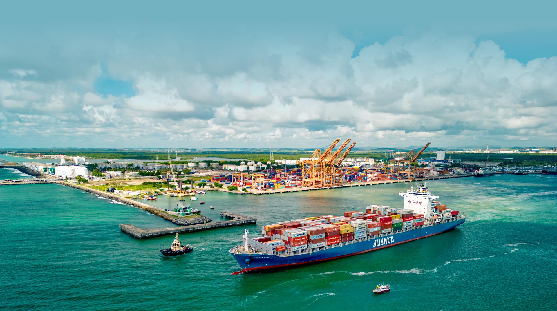 Uma foto aérea mostra um grande navio porta-contêineres azul e vermelho, carregado com contêineres coloridos, navegando em águas verde-esmeralda em primeiro plano. Ao fundo, um porto industrial está repleto de contêineres empilhados e várias grandes gruas amarelas sob um céu parcialmente nublado.