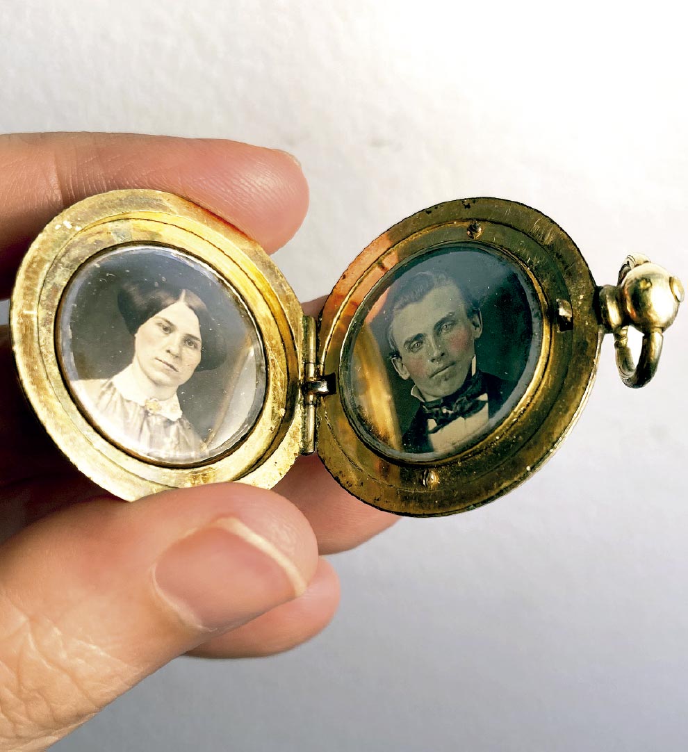 A imagem apresenta um plano detalhe (close-up) de uma mão segurando um pequeno relicário (medalhão) dourado e redondo, que está aberto, revelando dois retratos antigos em seu interior.

No lado esquerdo, vê-se a imagem de uma mulher de pele clara. Ela tem cabelos escuros, partidos ao meio e presos num coque baixo ou trança (estilo vitoriano), e veste uma roupa escura com uma gola de renda branca fechada por um broche dourado. Sua expressão é séria.

No lado direito, há o retrato de um homem jovem de pele clara, com as bochechas levemente rosadas (sugerindo uma colorização manual na fotografia). Ele tem cabelos curtos e escuros, e veste um paletó preto sobre uma camisa branca com uma gravata ou lenço escuro volumoso no pescoço.

O objeto tem uma borda metálica dourada com sinais de desgaste pelo tempo, uma dobradiça central e uma argola para corrente na extremidade direita. O fundo é branco e neutro.